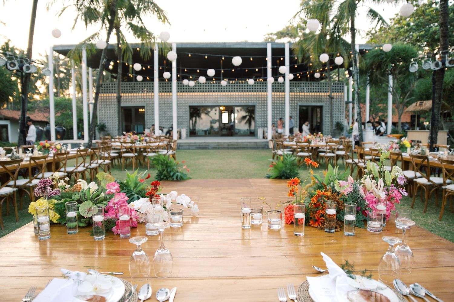 Elegant outdoor wedding reception with long wooden tables, tropical floral centerpieces, string lights, and white lanterns.