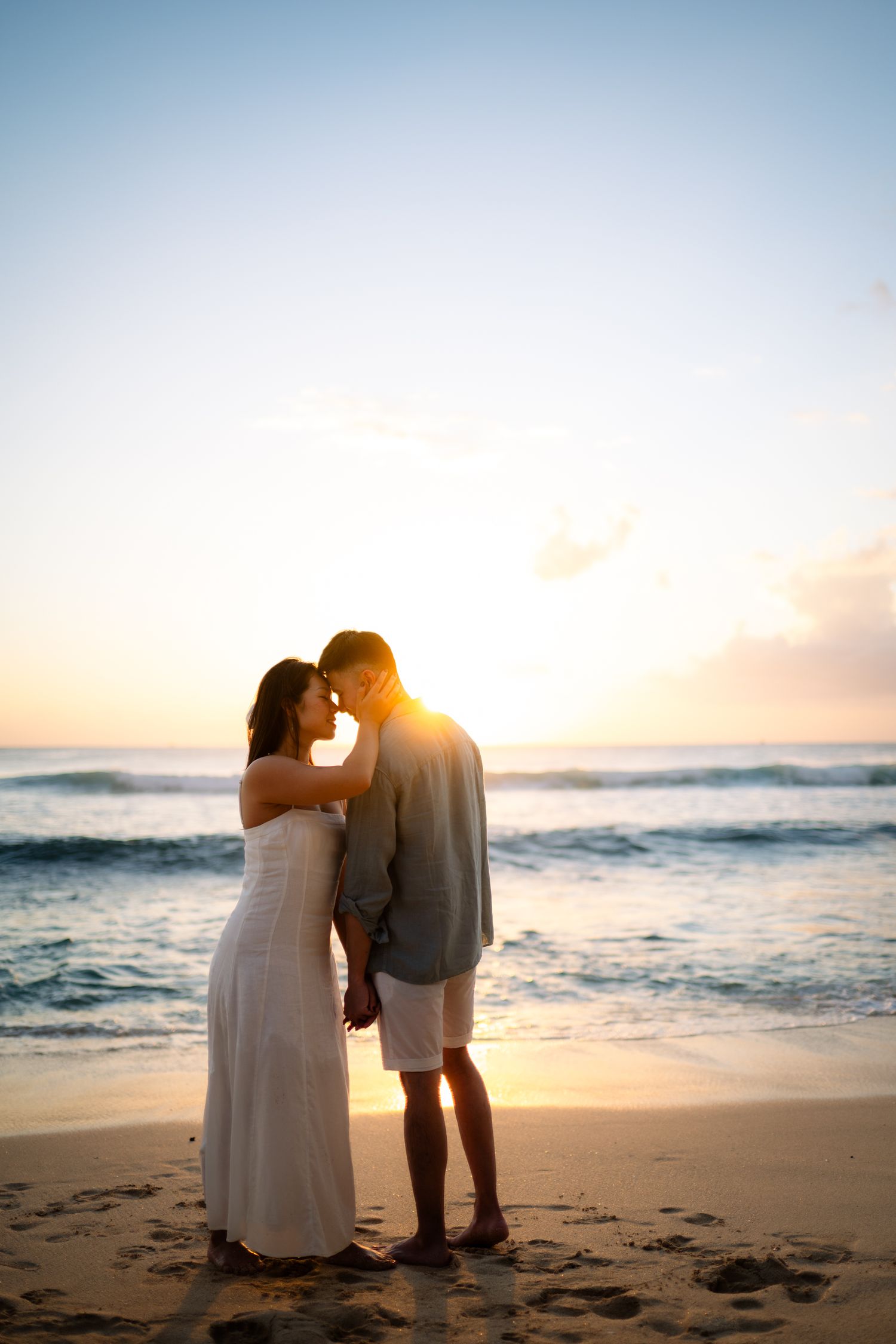 Romantic couple sharing a tender moment on a sandy beach at sunset, with ocean waves and golden sky behind them.