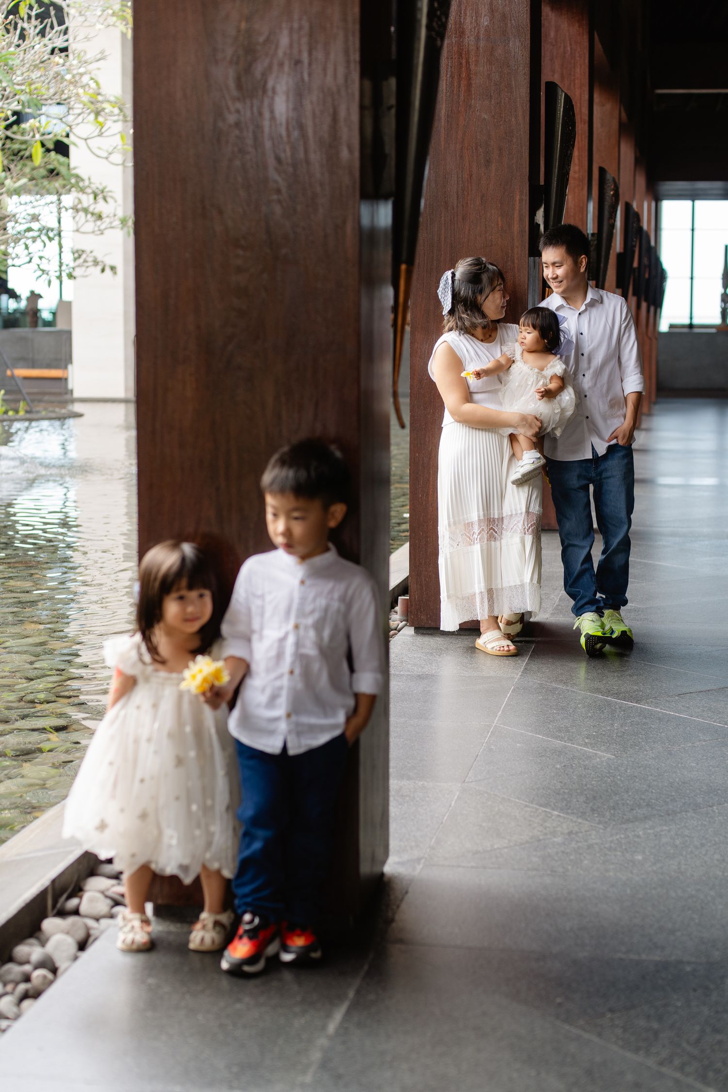 Asian family portrait with two children posing by wooden pillars while parents hold baby in modern interior.