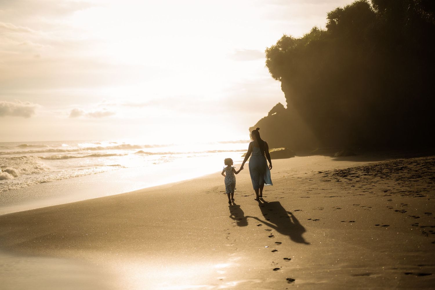 Mother and child walking hand in hand on a sandy beach at golden sunset with ocean waves and cliffs in background.