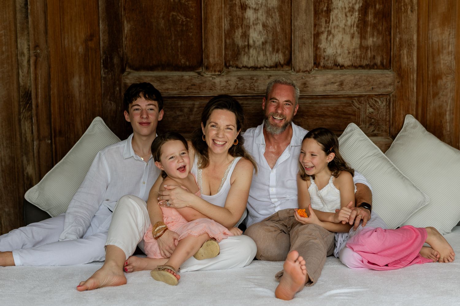 Happy family of five sitting together on a white bed, smiling warmly in a rustic wooden room.