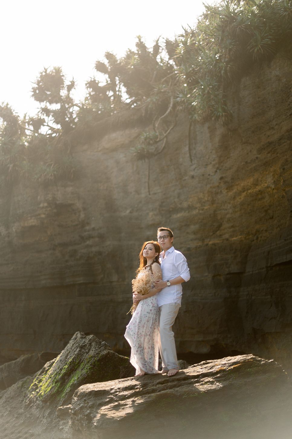 Couple embracing on rocky cliff at golden hour, woman in floral dress, man in white shirt, lush greenery backdrop.