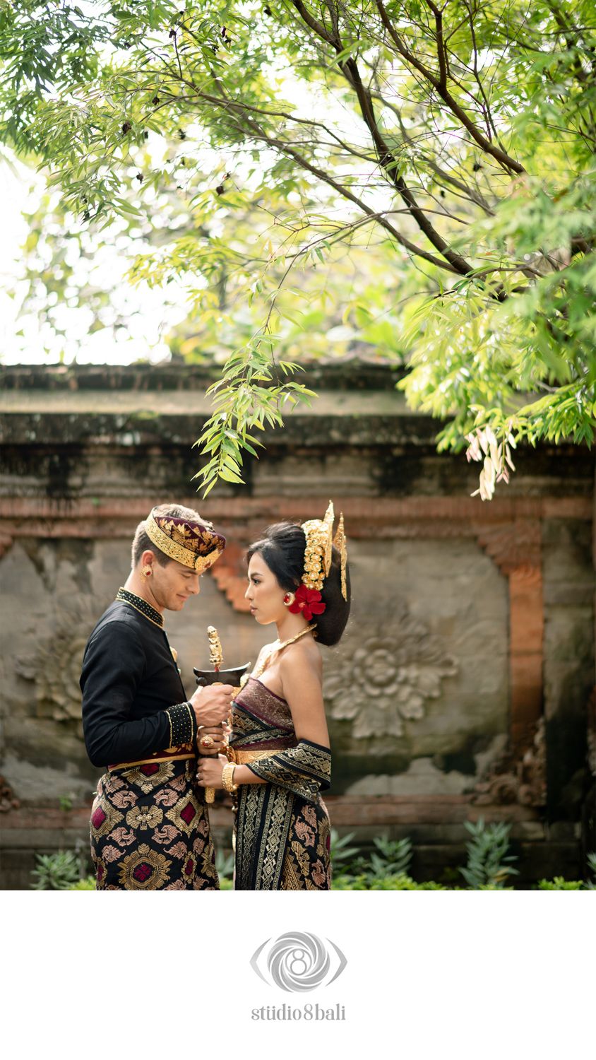 Balinese couple in traditional wedding dress sharing an intimate moment, surrounded by lush greenery and ancient stone walls.