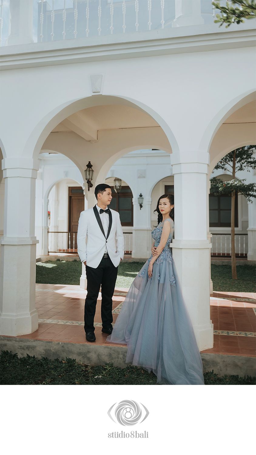 Couple in elegant attire posing under white arches at a colonial-style venue for a pre-wedding photoshoot.