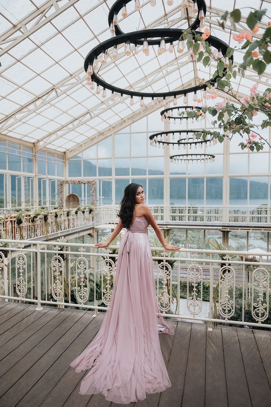 Woman in elegant pink strapless gown poses in a glass greenhouse with chandeliers and ornate white railings.