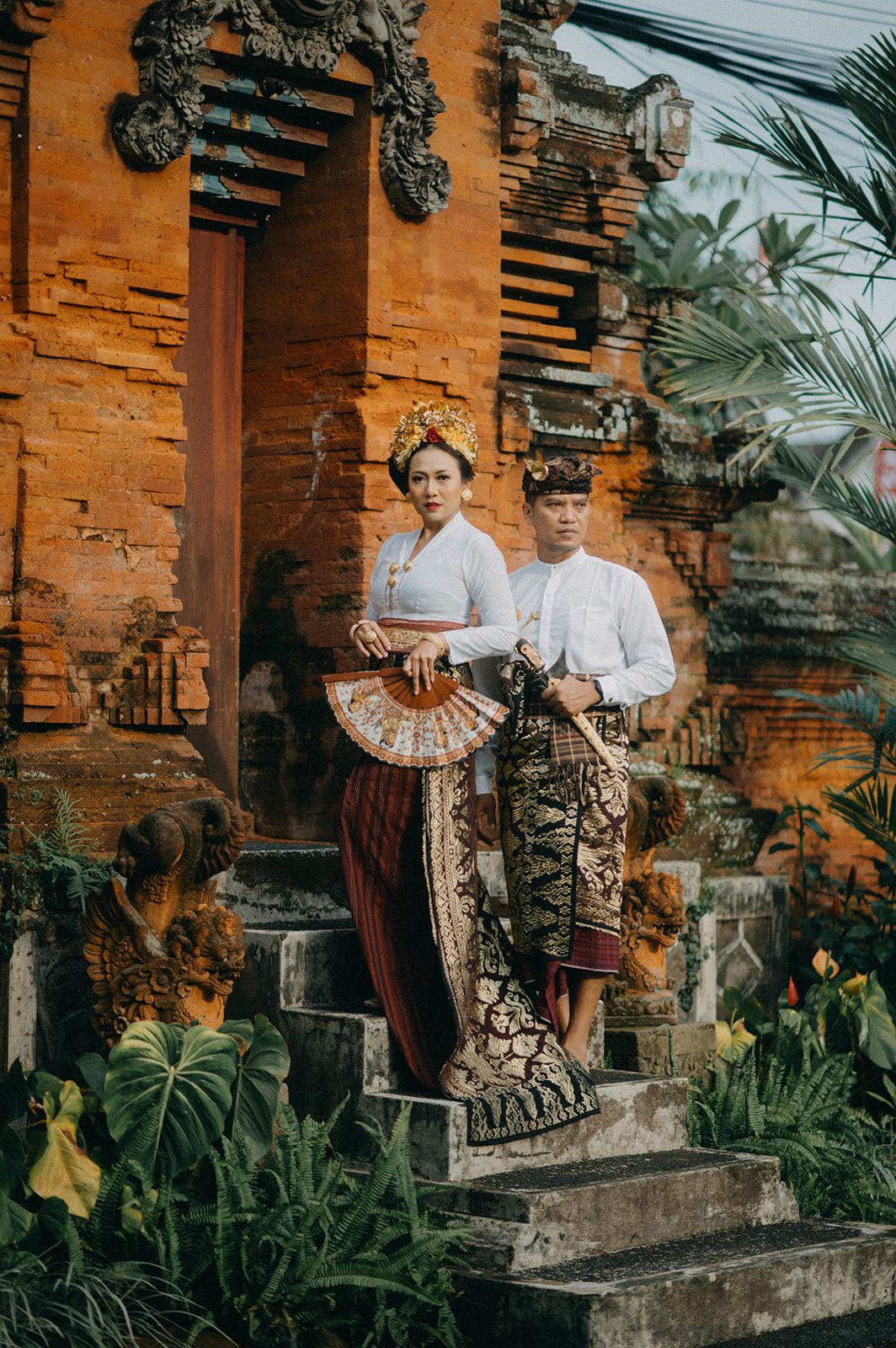 Balinese couple in traditional attire posing at an ornate temple gate surrounded by lush tropical greenery.