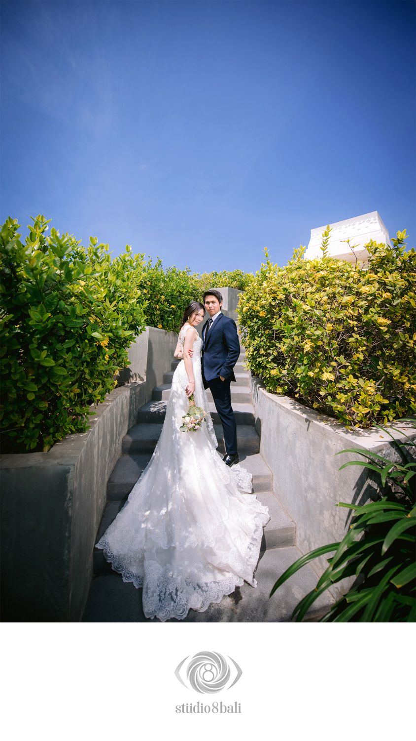 Bride and groom pose on outdoor stone steps surrounded by lush greenery under a bright blue sky.