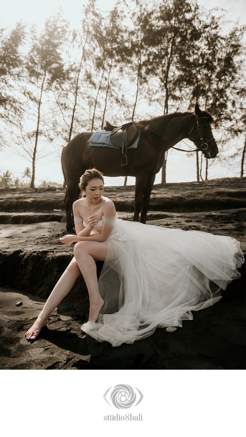 Bride in white tulle gown sits on rocks beside a saddled dark horse on a beach, in a romantic pre-wedding shoot.