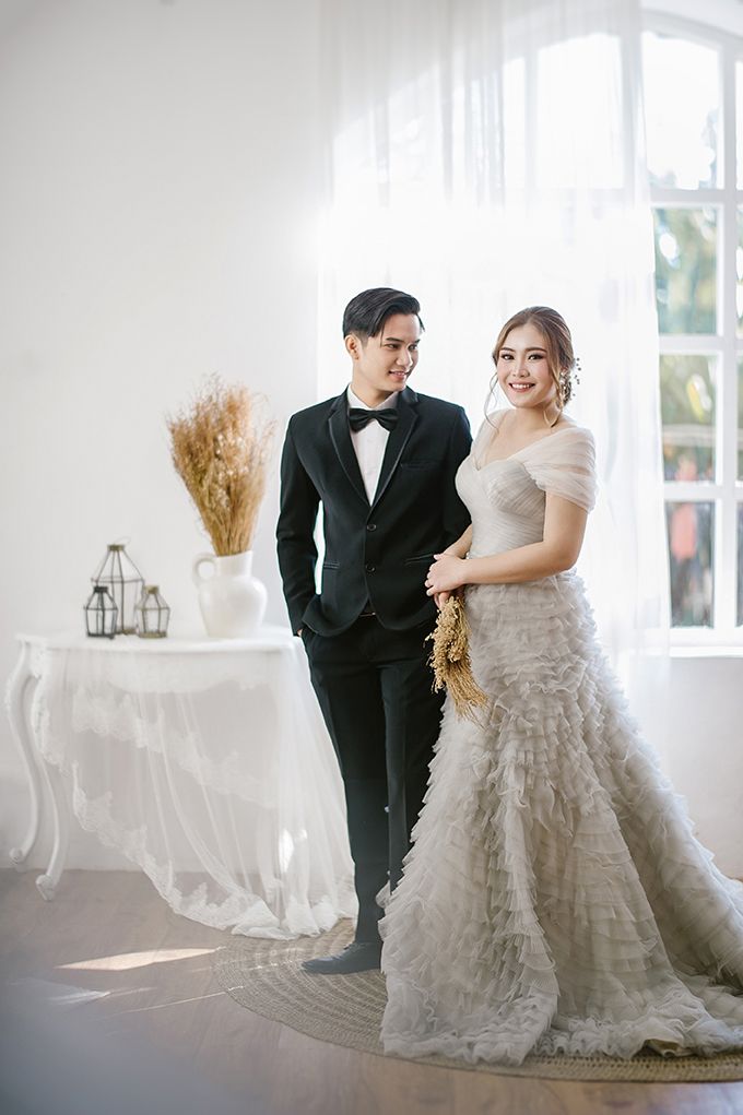 A couple poses in a bright studio, the groom in a black tuxedo and bride in a white ruffled gown holding dried flowers.