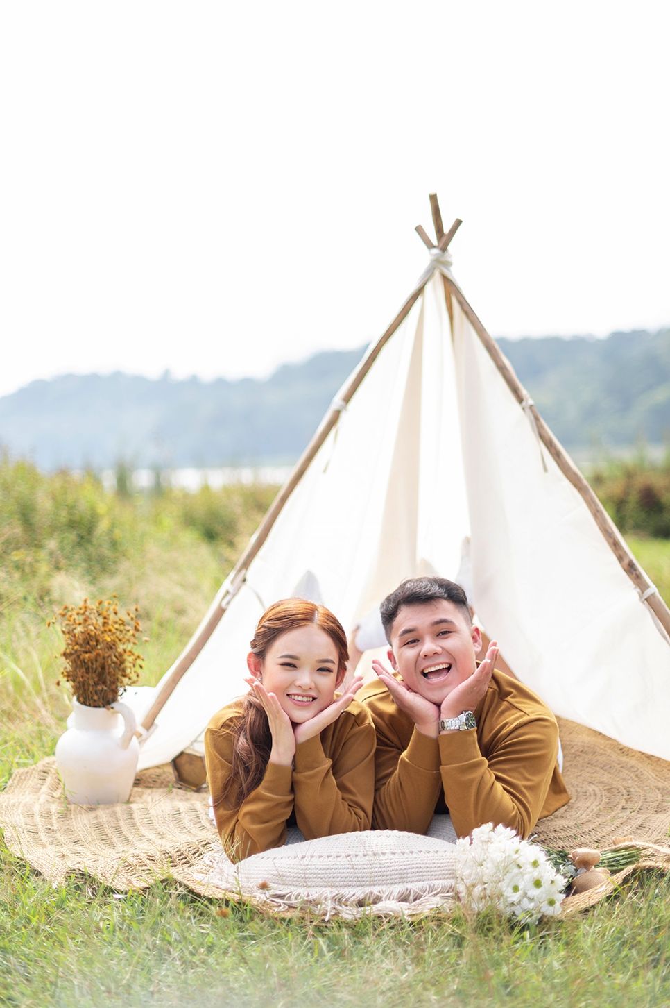 A smiling couple in matching brown outfits lying on a woven mat in front of a white teepee tent outdoors.