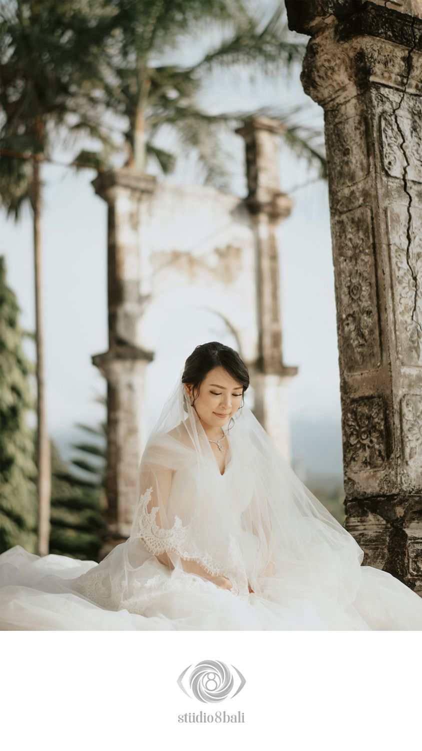 Bride in white wedding gown and veil seated among ancient stone columns in a serene Bali temple setting.