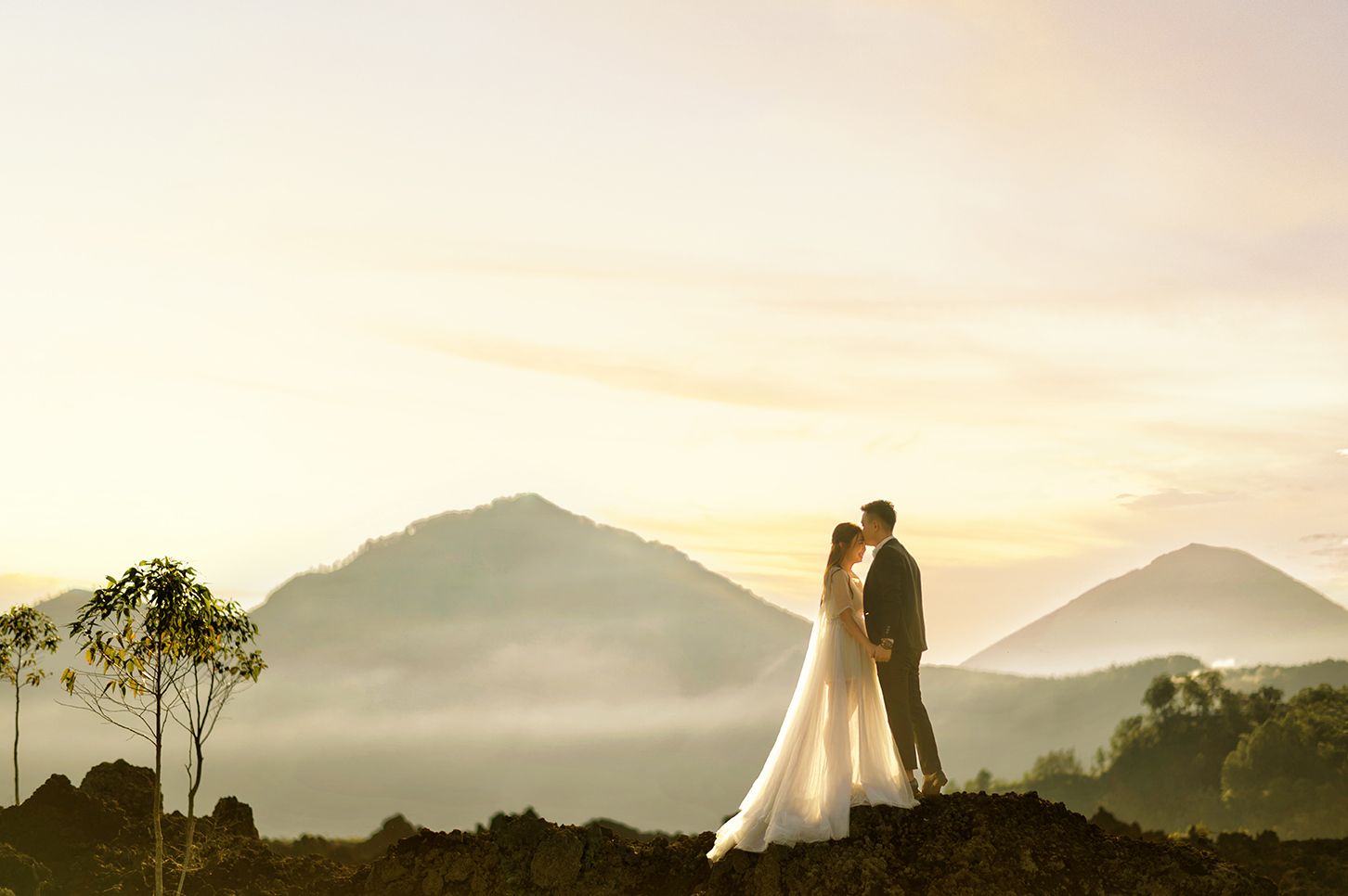 Romantic couple kissing on rocky hilltop with misty mountain peaks and golden sky backdrop.