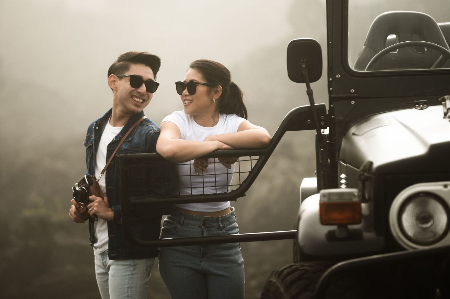A stylish couple in sunglasses leaning against a black jeep in a misty outdoor setting.