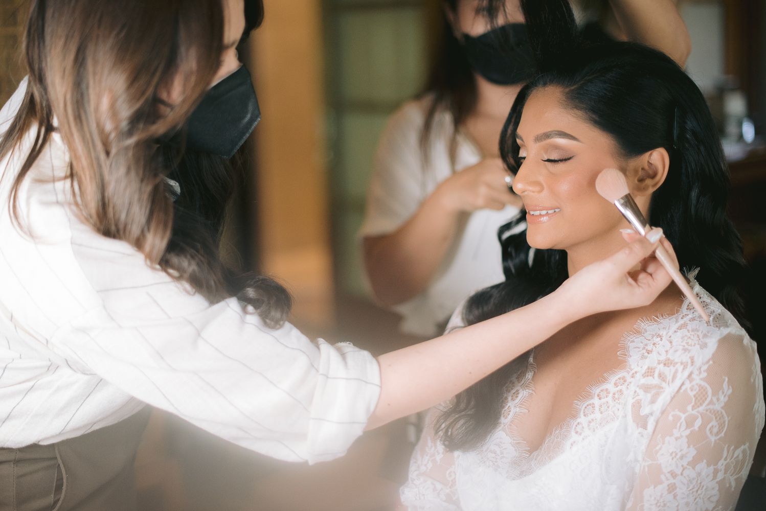 Makeup artist applying blush to a smiling bride in a lace dress during wedding preparations.