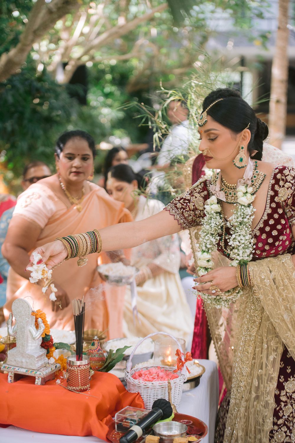 Bride in maroon bridal lehenga performing Hindu wedding rituals at decorated ceremony table with flowers and offerings.