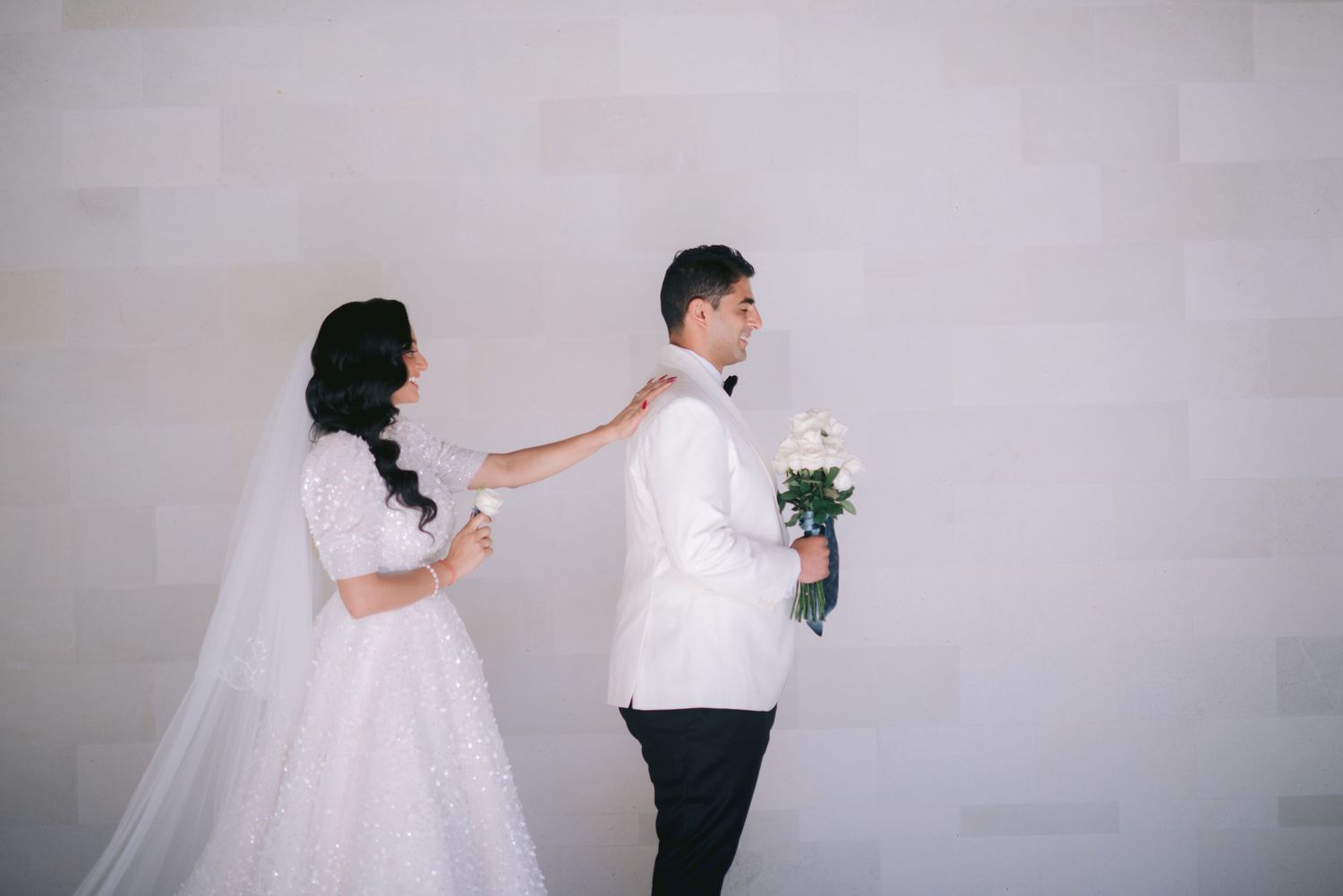 Bride in white lace gown adjusts groom's white tuxedo jacket as he holds a bouquet against a neutral wall.