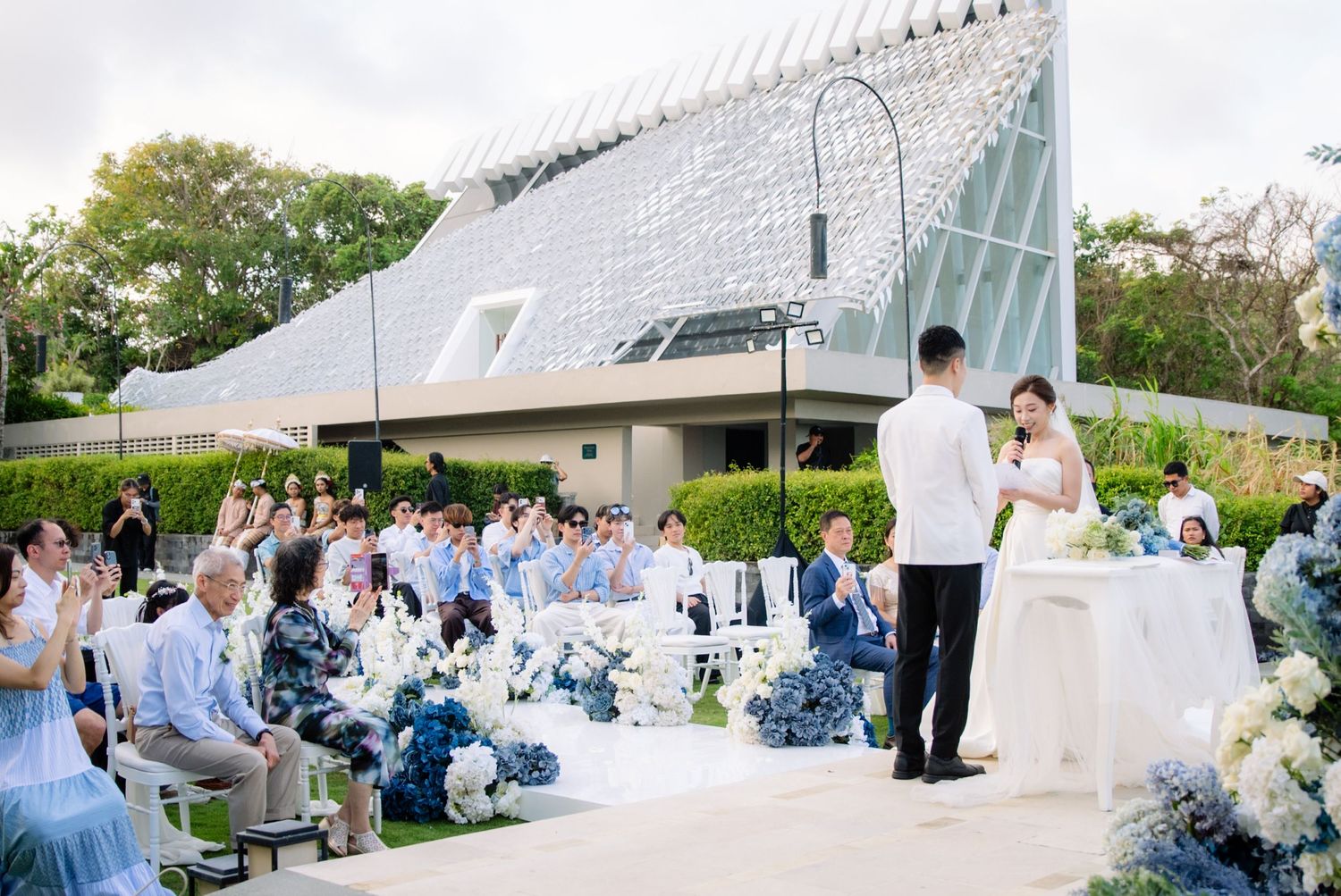 Bride and groom exchange vows at an outdoor garden wedding ceremony with seated guests, blue and white flowers, and a modern glass chapel be