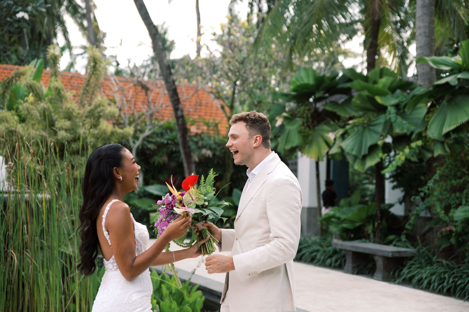 Bride in white dress receives tropical bouquet from groom in ivory suit outdoors at lush garden venue.