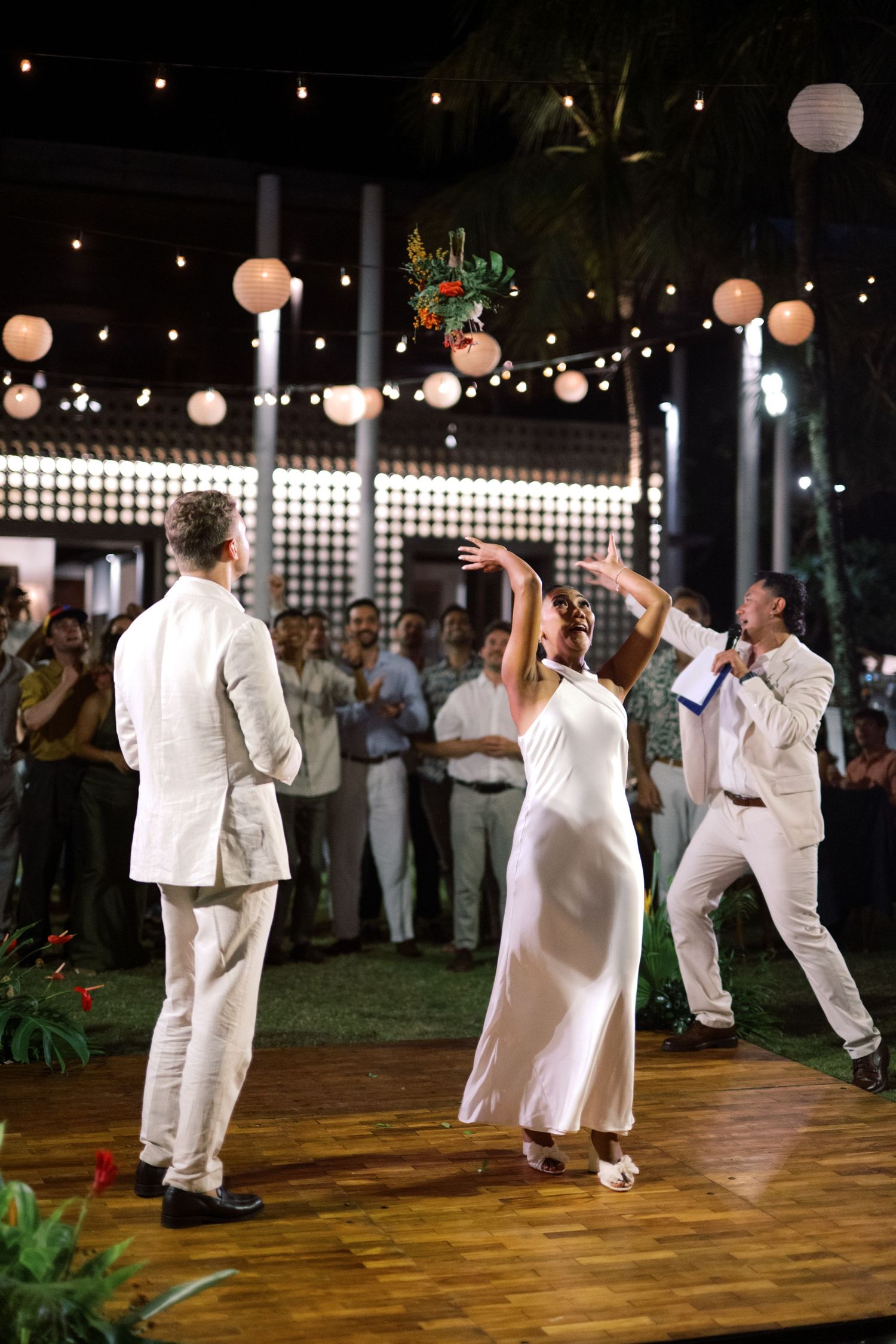 Bride tosses tropical bouquet to excited guests on dance floor at outdoor wedding reception with string lights.