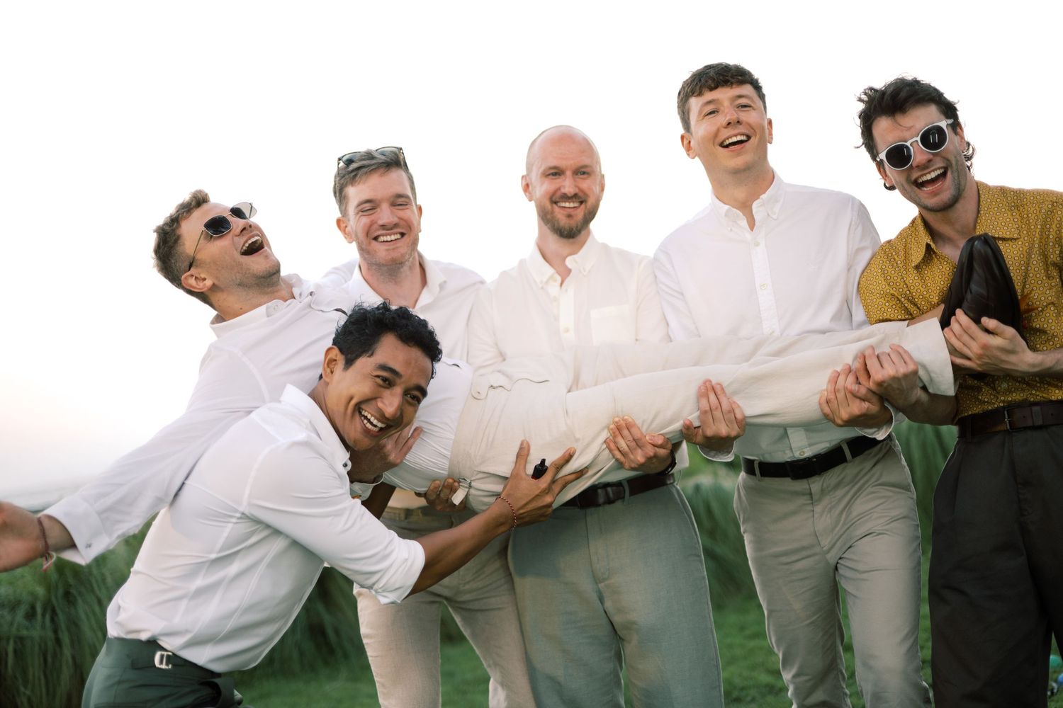 Group of groomsmen laughing and lifting the groom outdoors at a tropical wedding celebration.