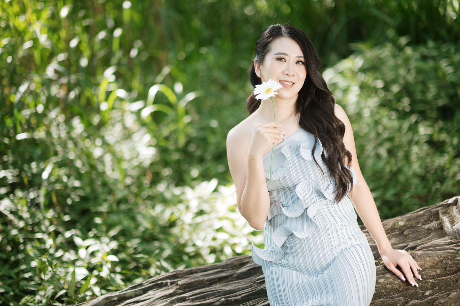 Young Asian woman in light blue dress holding white flower, smiling outdoors among lush green foliage.