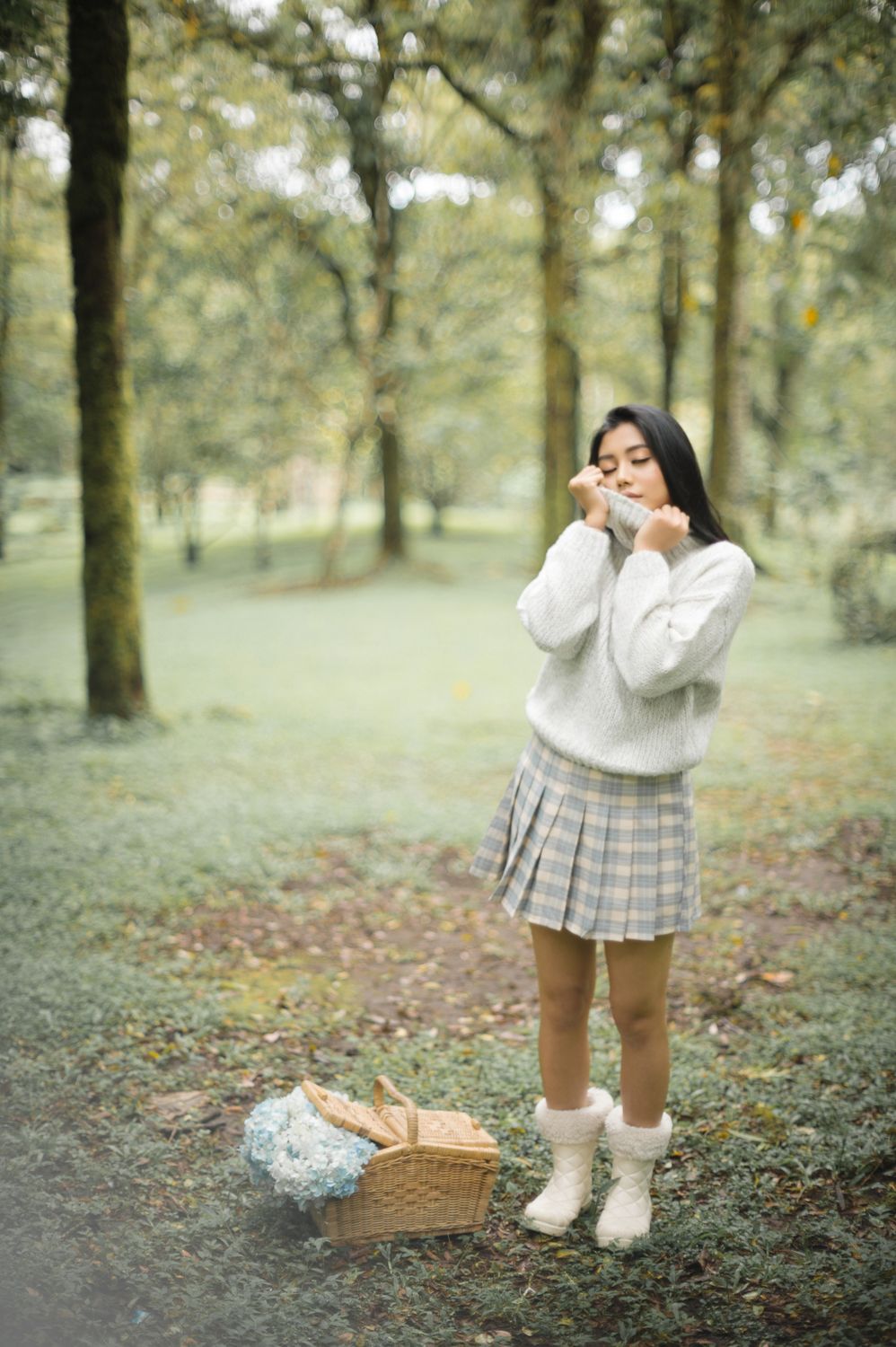 Young woman in white sweater and plaid skirt stands in a misty forest park beside a wicker picnic basket.