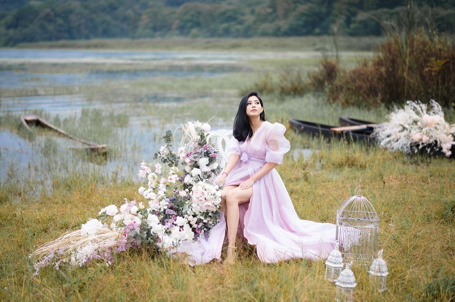 A woman in a pink gown poses by a lake surrounded by flowers and lanterns in a dreamy outdoor photoshoot.