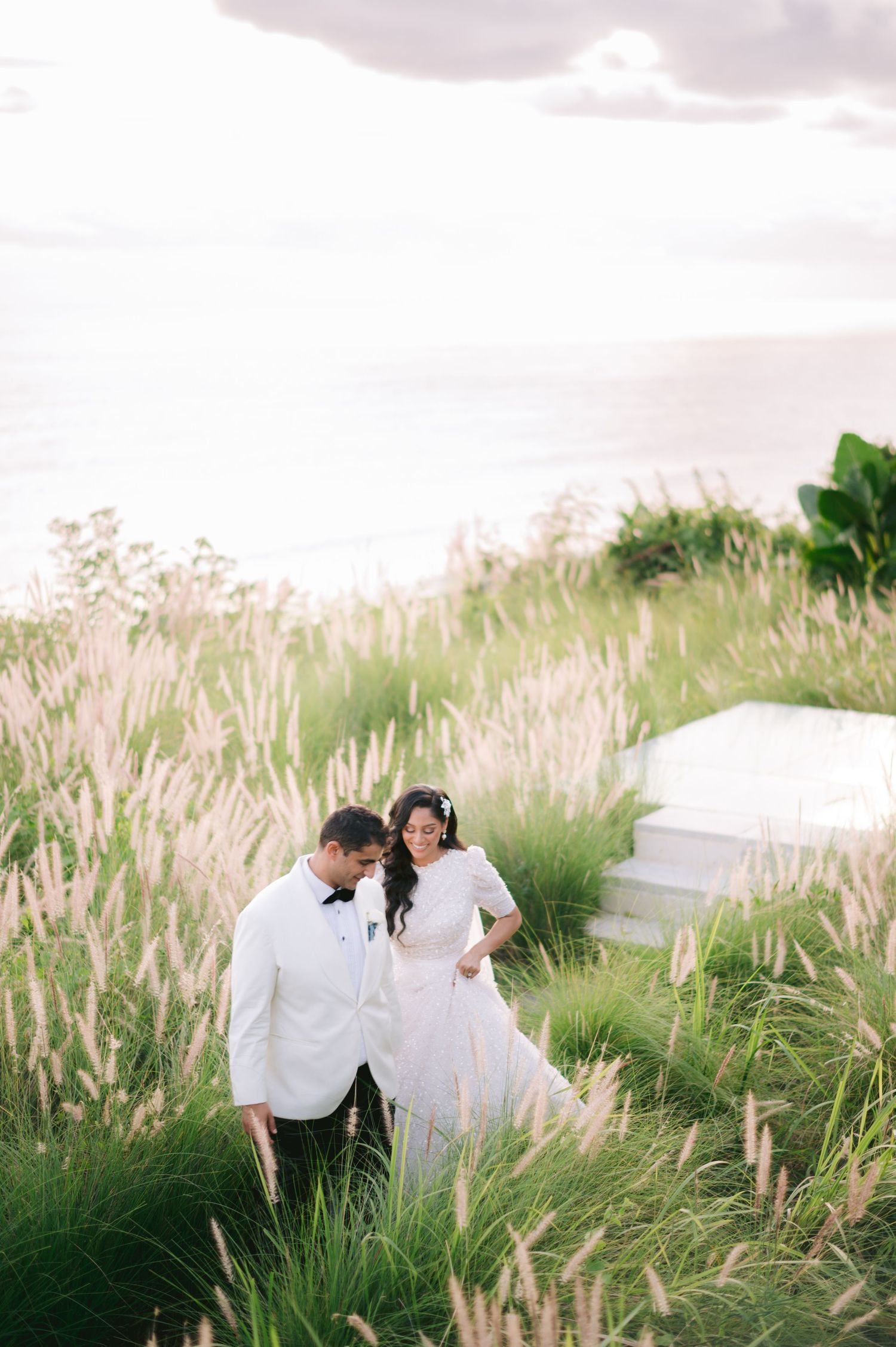 A couple in wedding attire walks through tall grass with an ocean view at sunset.