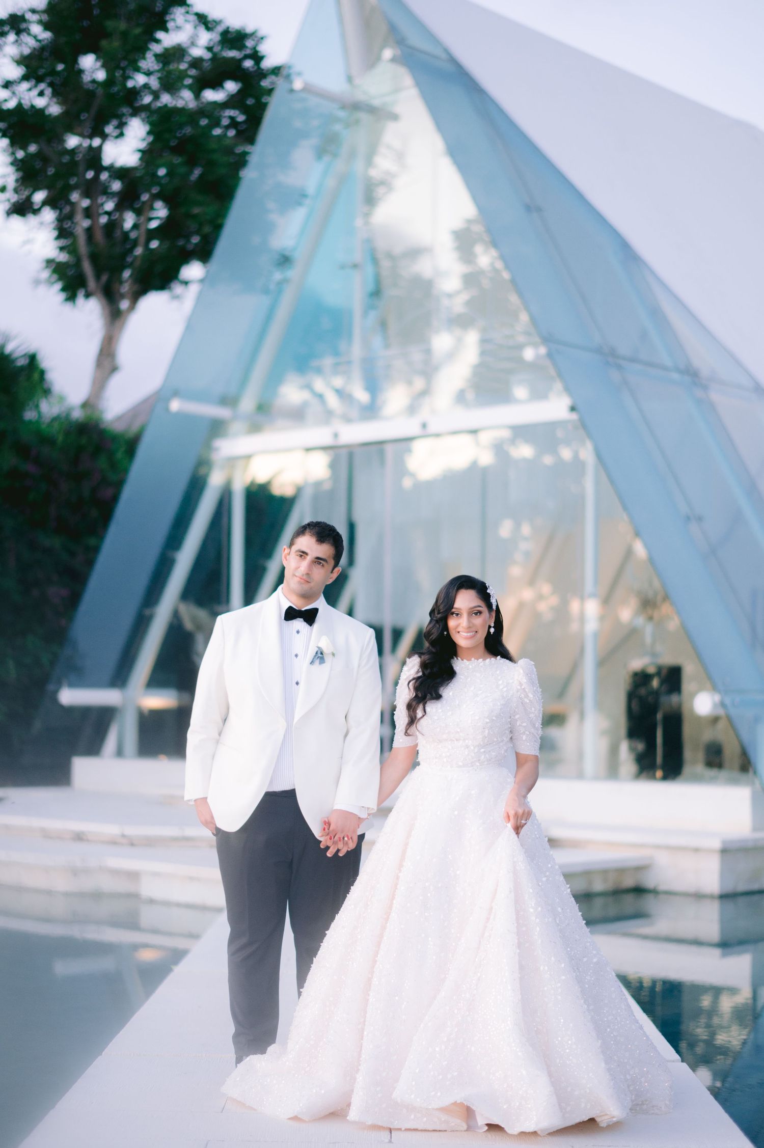 Bride and groom hold hands in front of a modern glass A-frame chapel, wearing elegant white wedding attire.