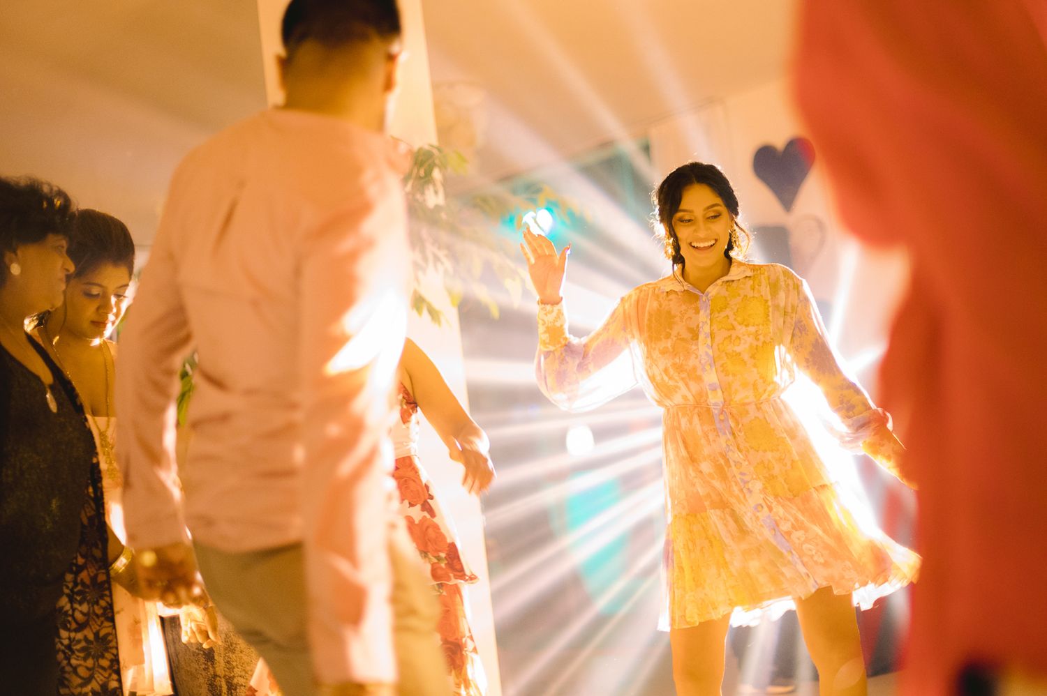 Woman in yellow floral dress dancing joyfully at a vibrant celebration with colorful lights.