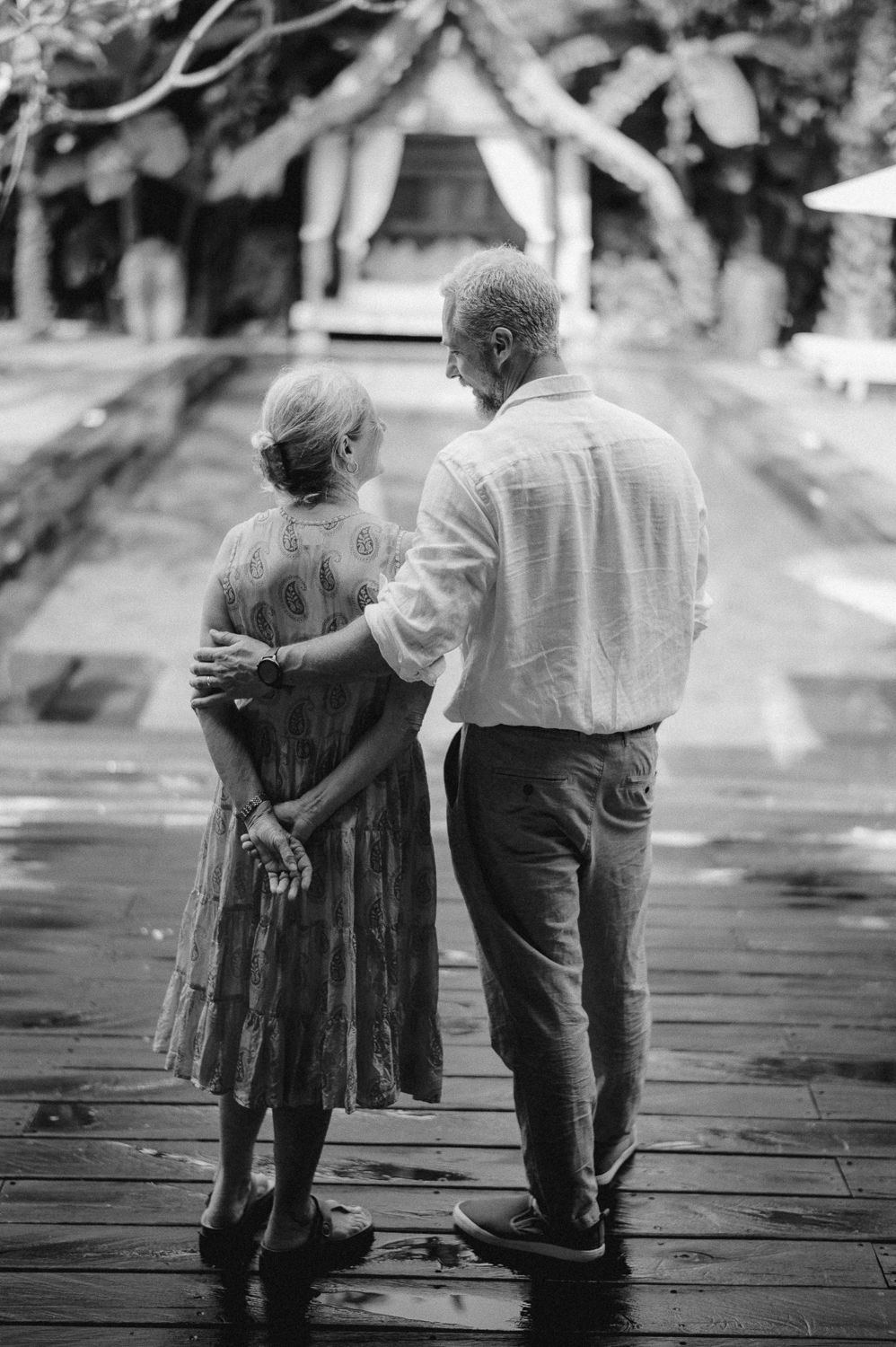 Black and white photo of an elderly couple walking hand in hand on a wooden deck near a garden.