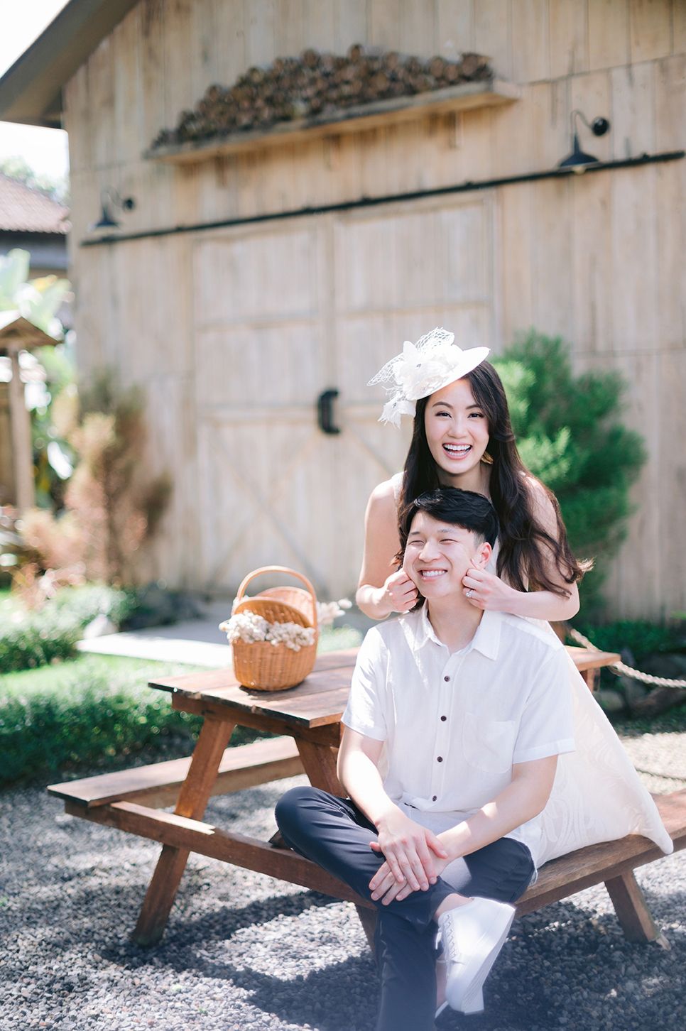Joyful woman in white fascinator covering seated man's eyes at a rustic outdoor barn setting.