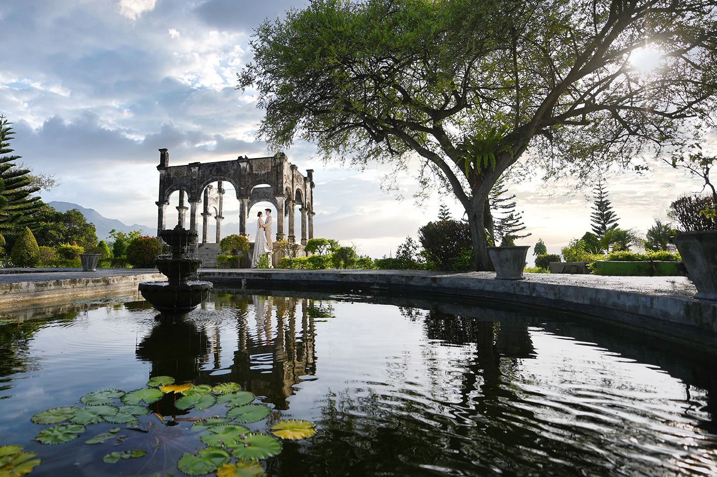 Serene garden pond with lily pads reflecting a stone archway monument and large tree at sunset.