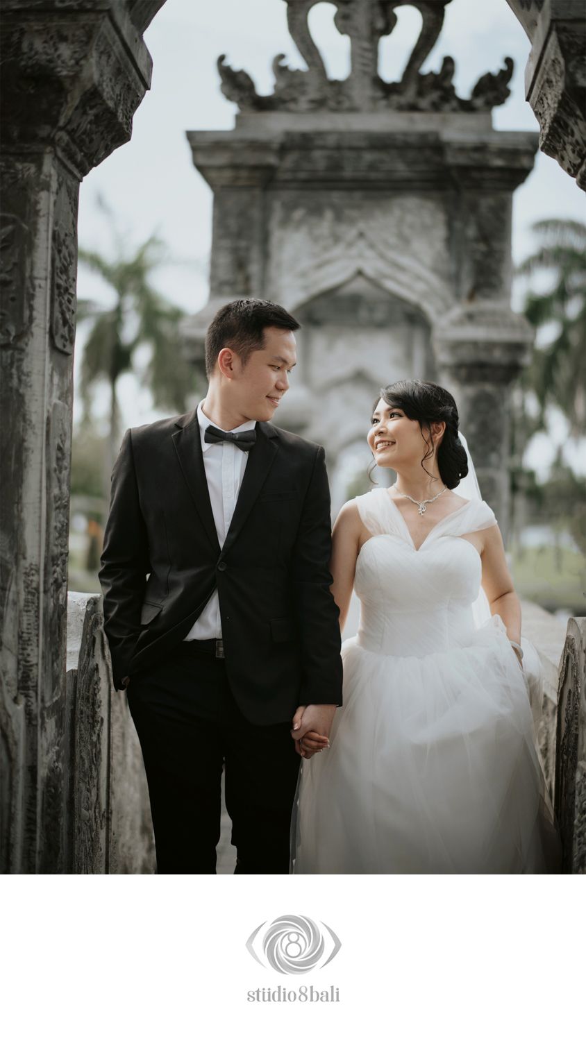 Couple in wedding attire holding hands at a historic stone monument, groom in black tuxedo, bride in white gown, smiling.