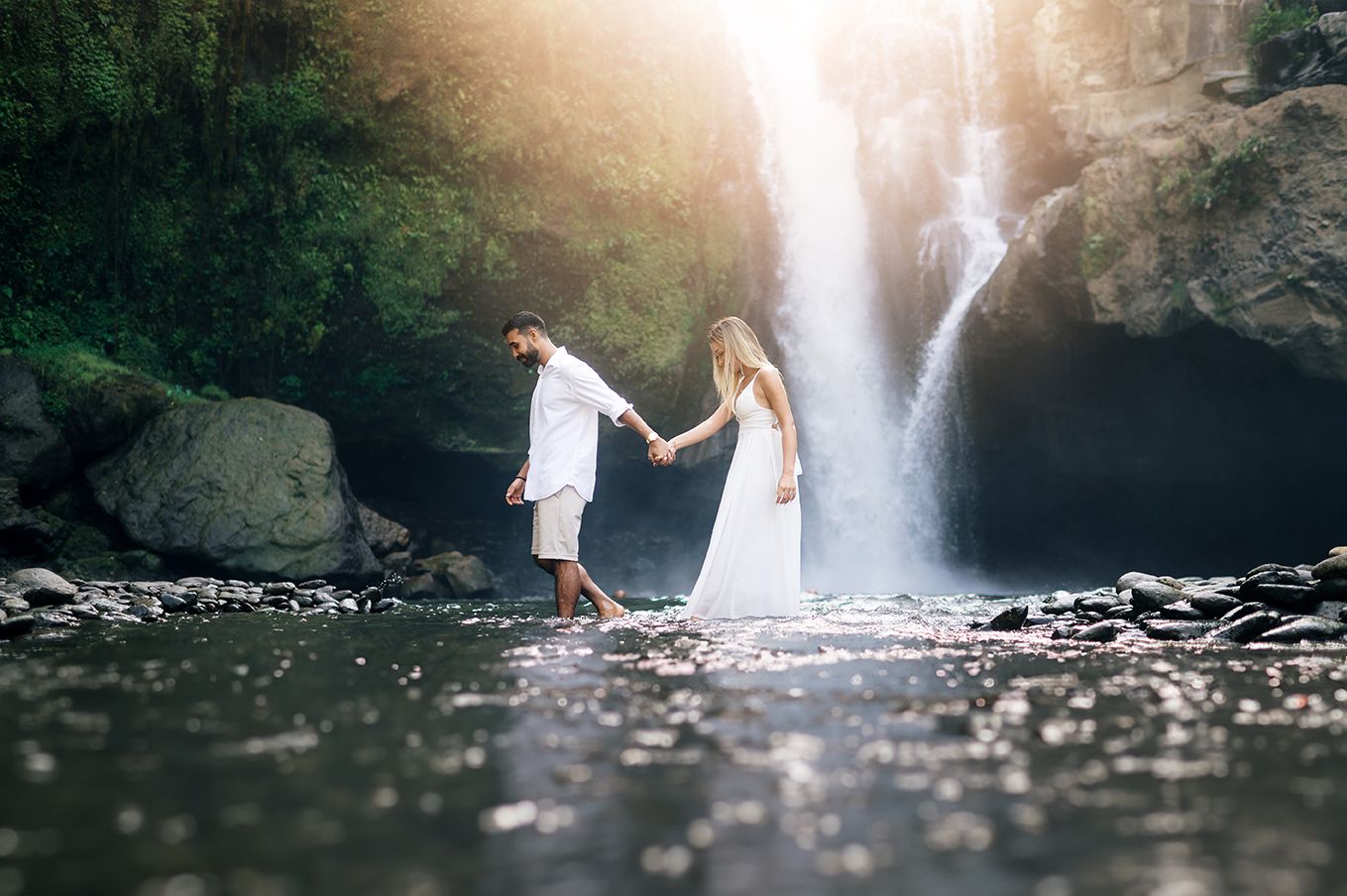 Couple in white holding hands by a serene waterfall surrounded by lush greenery and rocks.