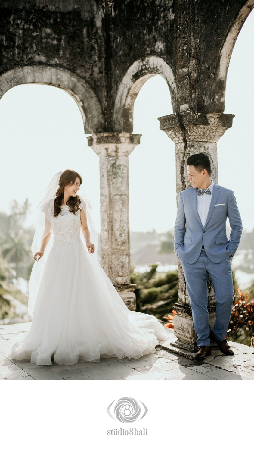 Bride in white gown and groom in blue suit pose under stone arches at a scenic Bali wedding venue.
