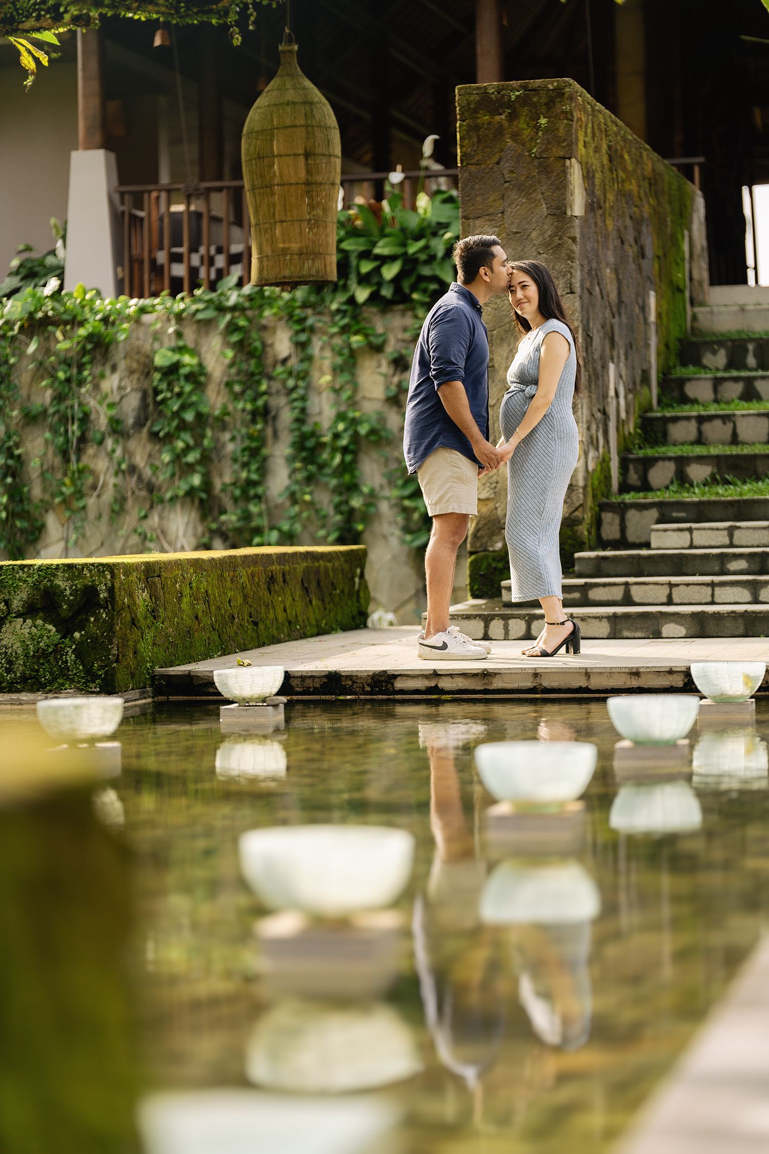 Couple sharing a romantic moment by a serene garden pond, surrounded by lush greenery and stone steps Maya Ubud Resort and Spa in Bali.