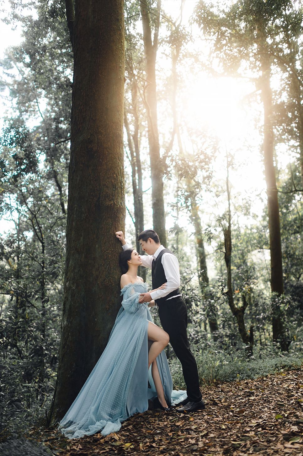 Couple embracing by a large tree in a sunlit forest, woman in a flowing blue gown, man in formal attire.