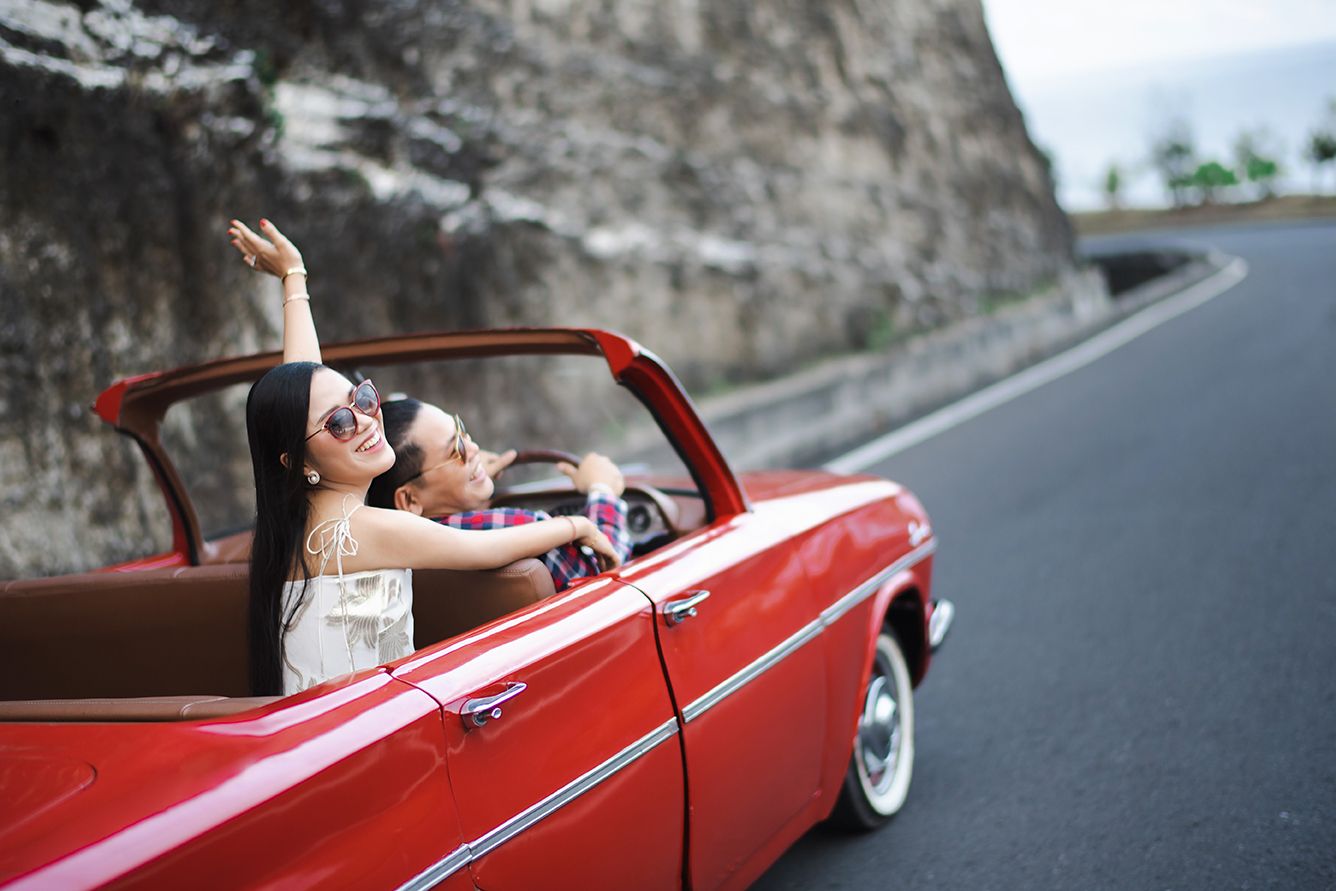 Two young women enjoy a scenic road trip in a classic red convertible car, one raising her arm joyfully.