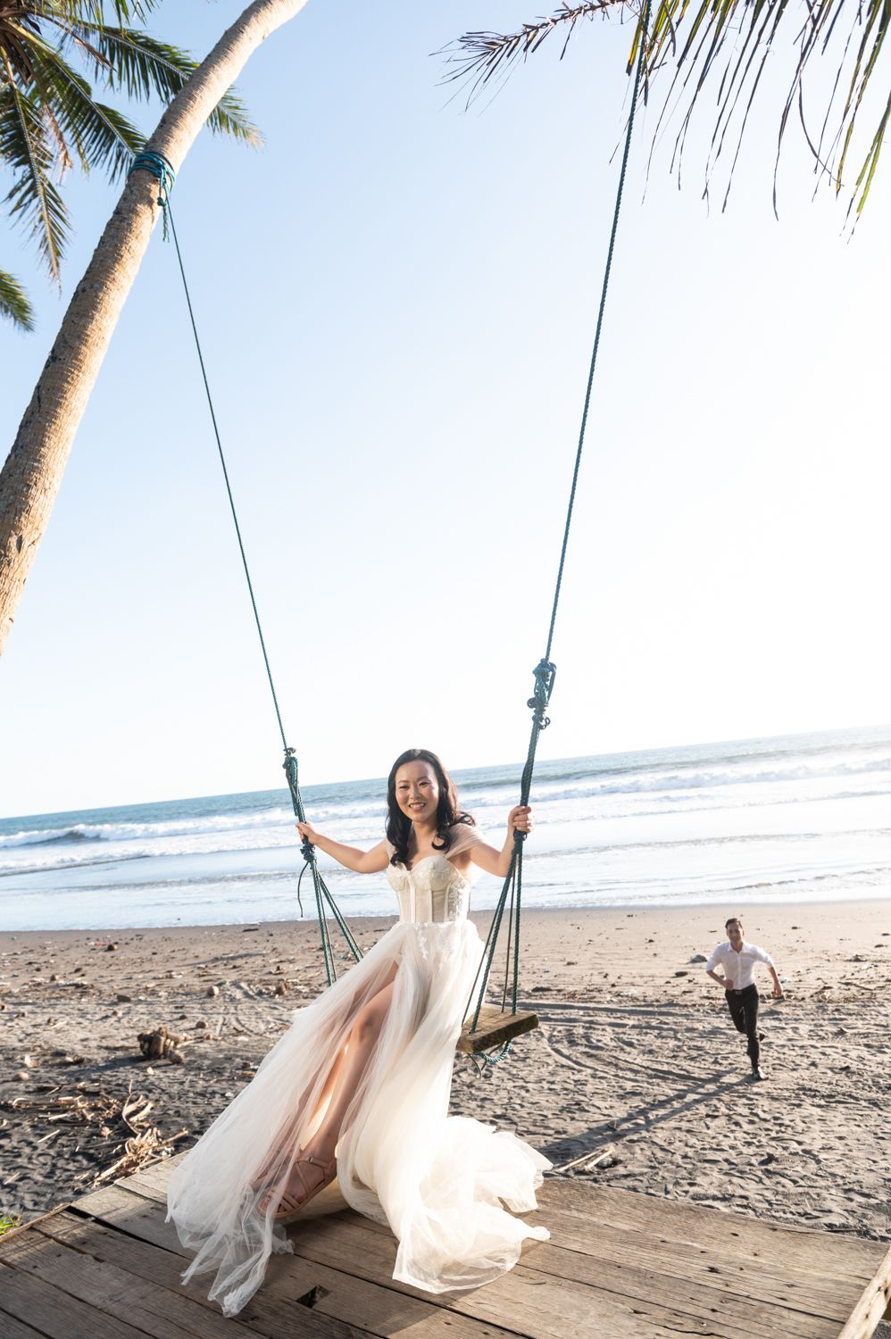 Bride in white gown sits on beach swing smiling, groom running toward her with ocean and palm trees in background.