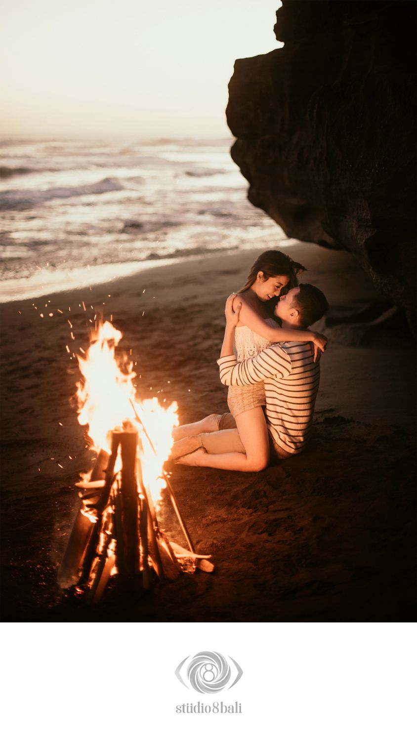 Couple embracing romantically by a beach bonfire at sunset, with ocean waves and rocky cliffs in the background.