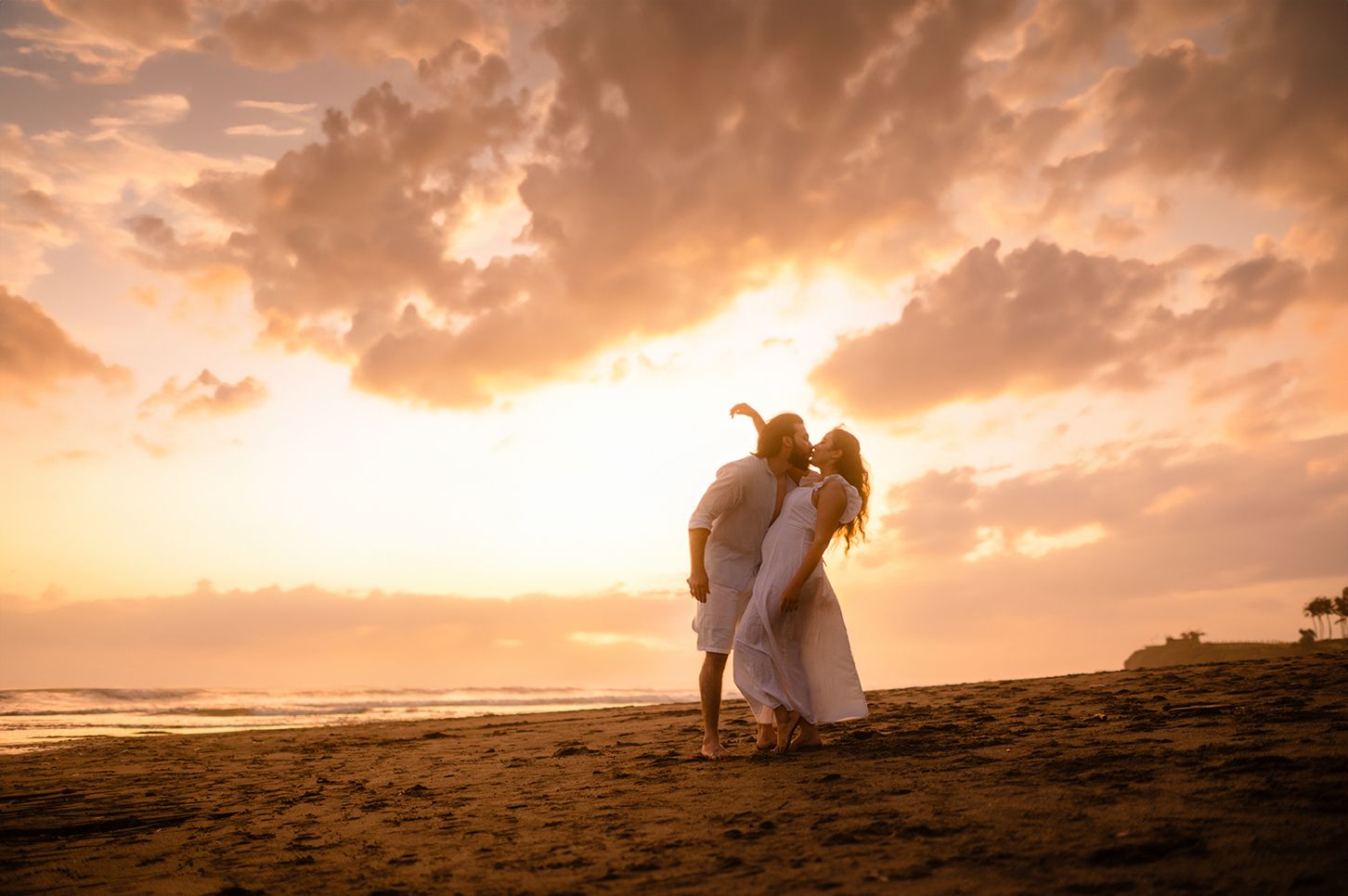 Couple embracing on a sandy beach at golden sunset with dramatic cloudy sky and ocean waves.