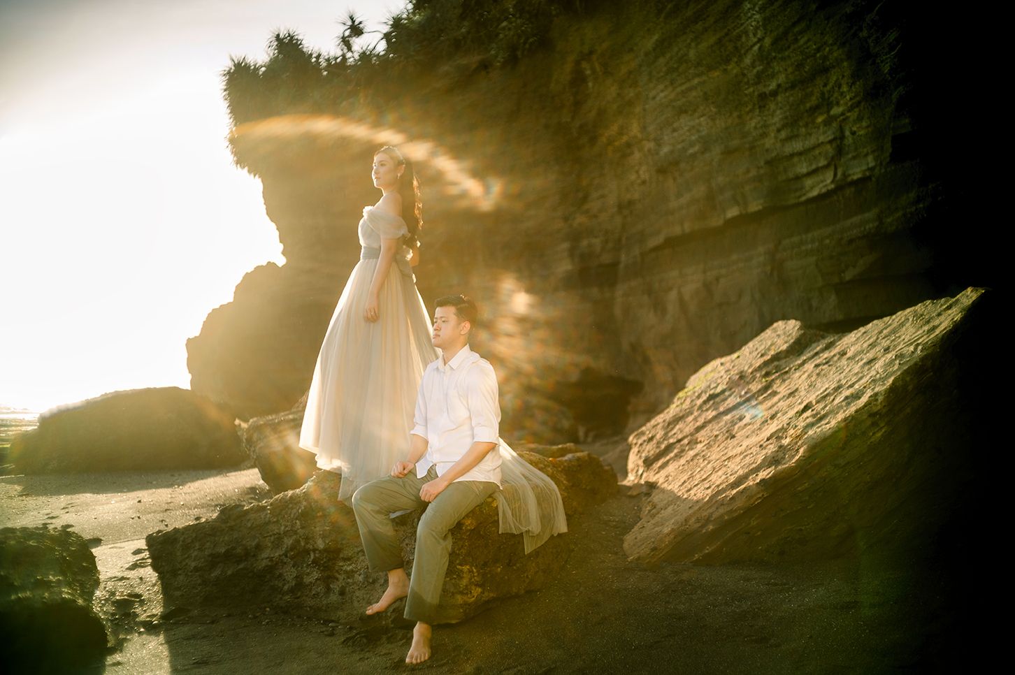 Couple in wedding attire posing on rocky coastal cliffs with golden sunlight streaming behind them.