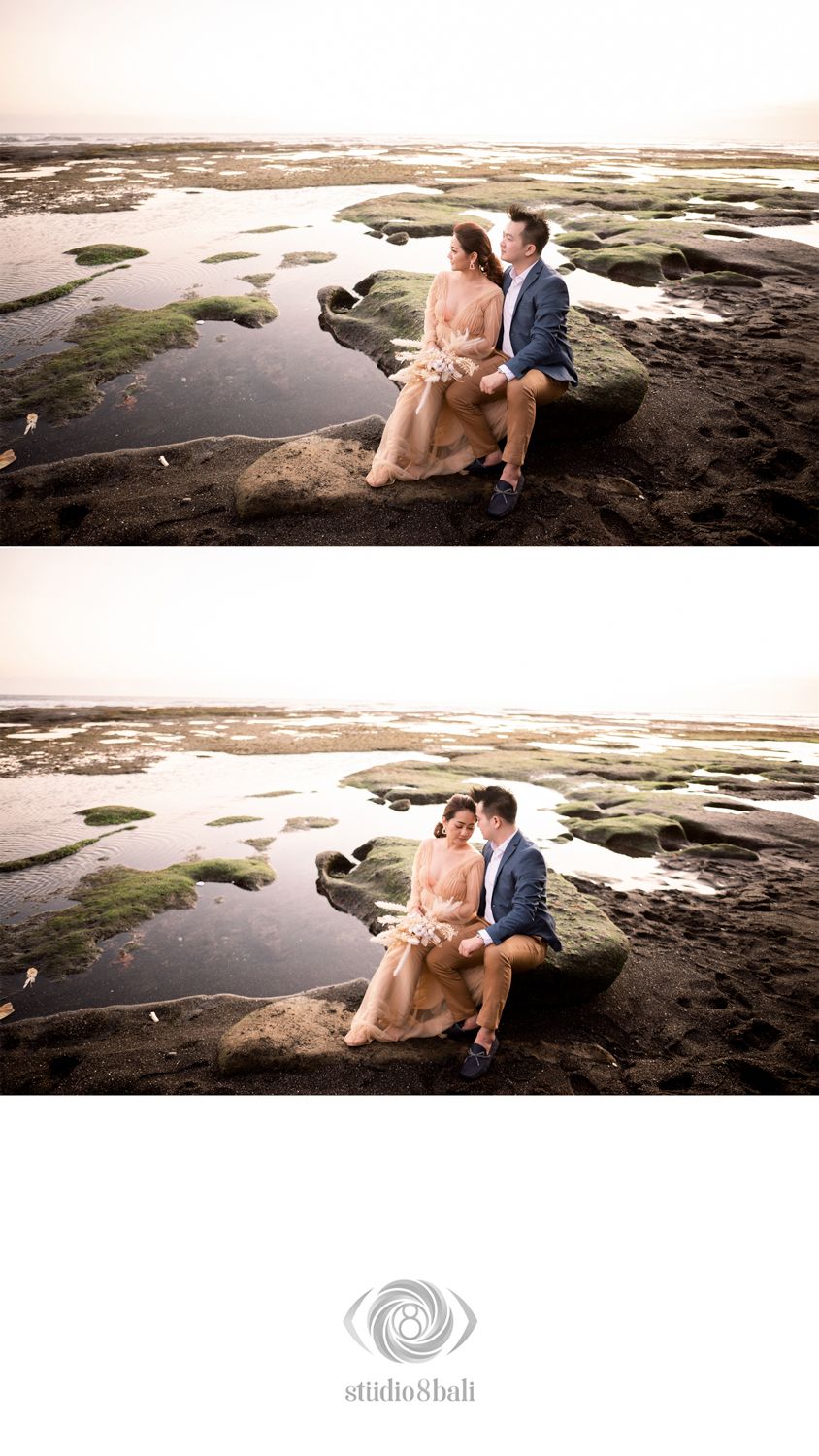 Couple sitting on rocky beach at sunset, woman in floral dress and man in blue suit, romantic pre-wedding photo by Studio 8 Bali.