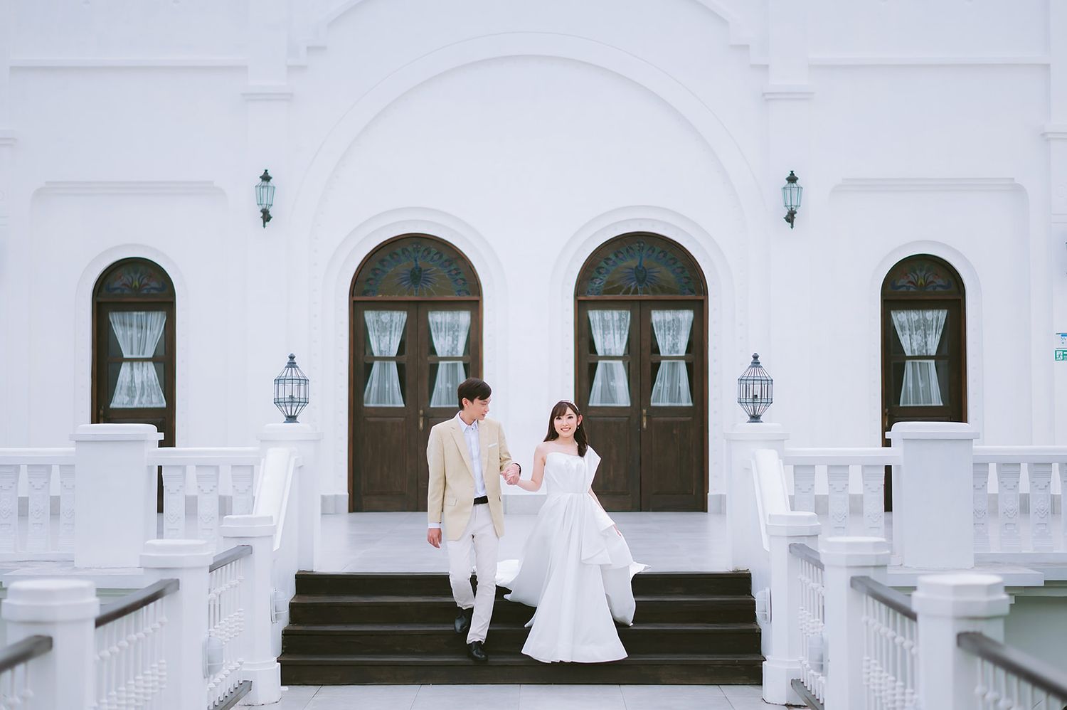 Couple in wedding attire posing on white steps of elegant building with arched doors and windows.