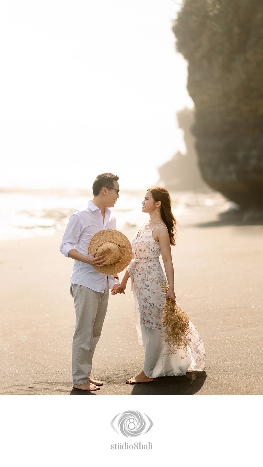 Couple holding hands on a sunlit beach in Bali, man holding straw hat, woman in floral dress with dried flowers.