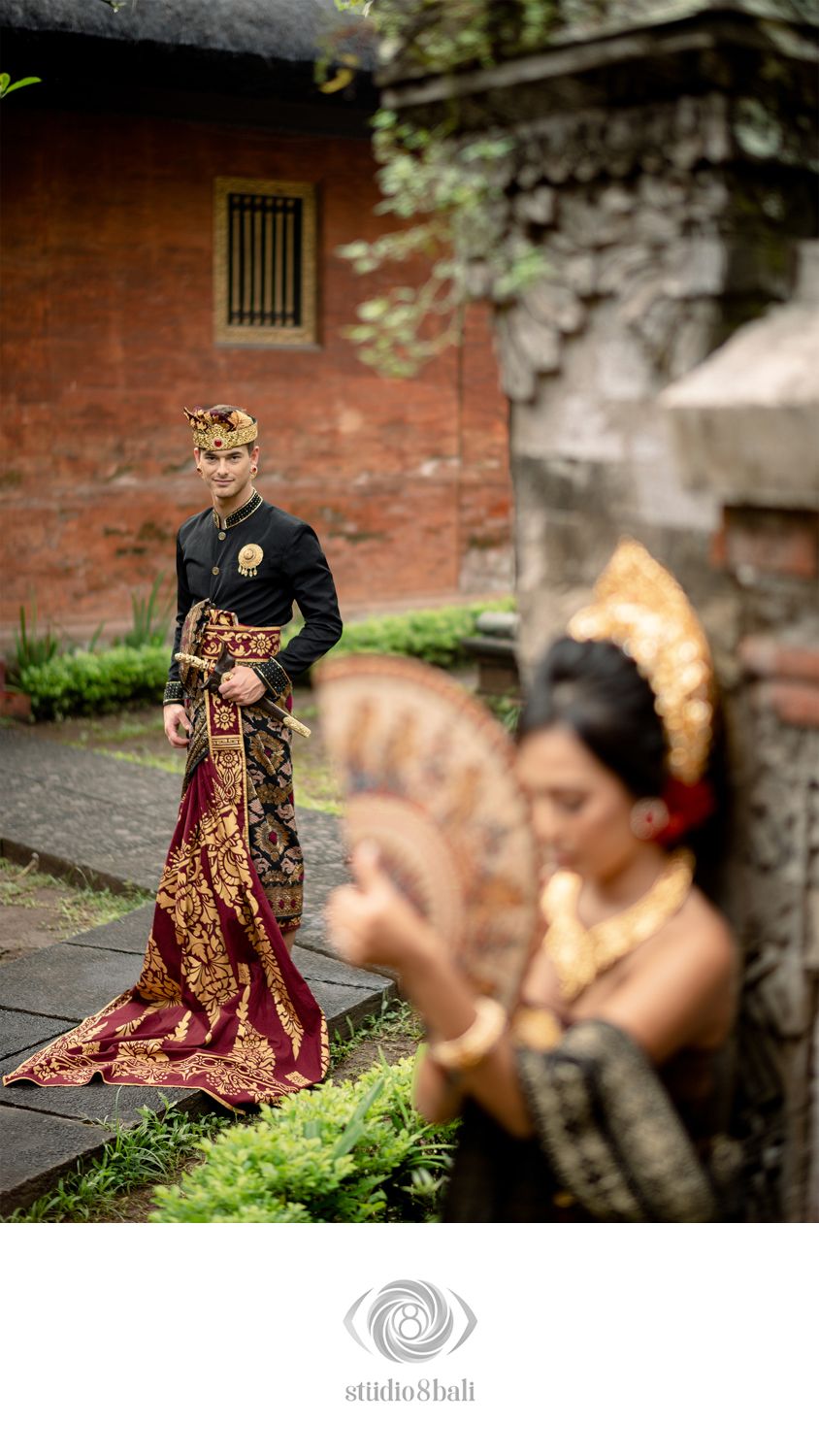 Balinese couple in traditional wedding attire, groom in black and gold approaching bride holding decorative fan near stone wall.
