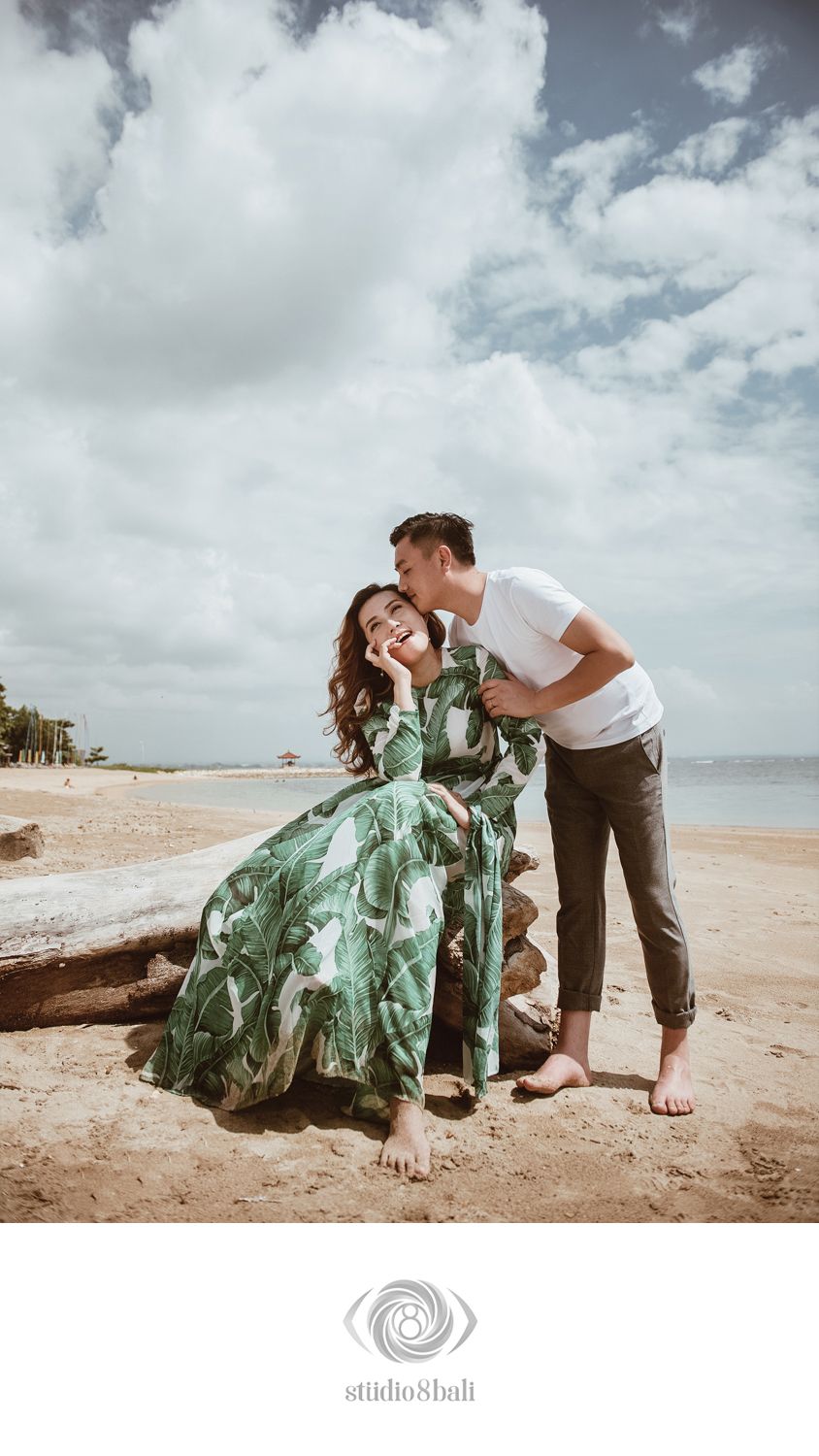 Couple sharing a romantic moment on a sandy beach, woman in green floral dress laughing as man kisses her cheek.