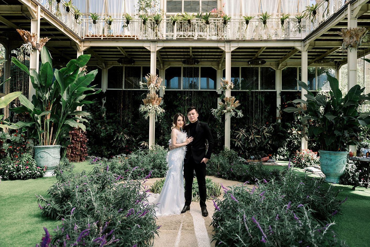 Couple in wedding attire posing in lush garden courtyard of elegant venue with tropical plants and balcony.