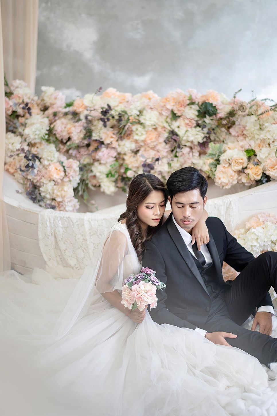 Elegant couple in wedding attire sitting together before a lush floral backdrop, bride holding a soft pink bouquet.